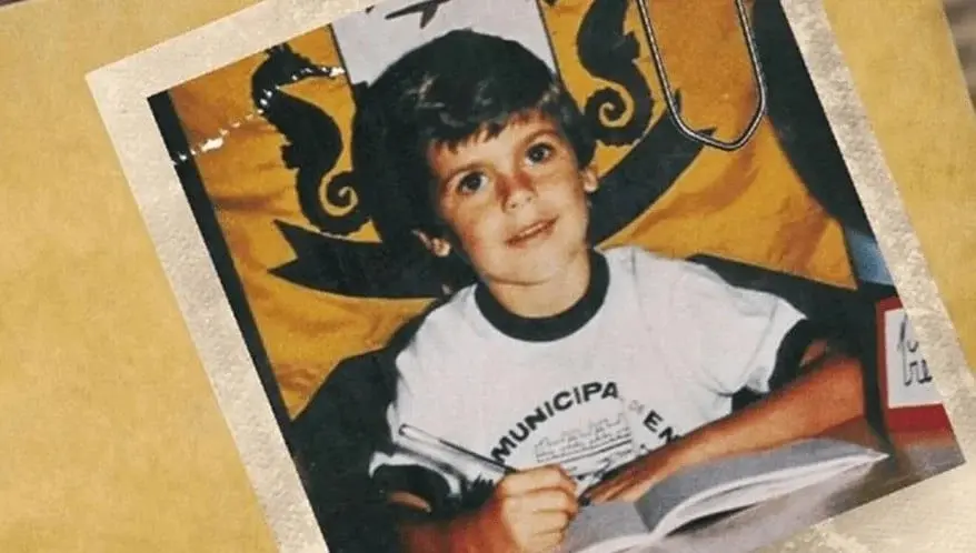 Uma fotografia antiga e levemente desgastada mostra o menino Evabdri sorrindo enquanto escreve em um caderno, vestindo uma camiseta branca de uniforme escolar. A imagem está presa por um clipe de papel a uma pasta de cor parda, com um brasão amarelado ao fundo que apresenta figuras de cavalos-marinhos.