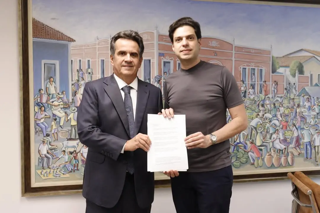 Dois homens posam lado a lado em um ambiente interno, segurando juntos um documento voltado para a câmera. O homem à esquerda veste terno escuro, camisa branca e gravata; o da direita usa camiseta escura e relógio. Ao fundo, há um grande quadro colorido retratando uma cena urbana com várias pessoas, barracas e atividades cotidianas. À direita da imagem, parte de uma cadeira de madeira é visível.