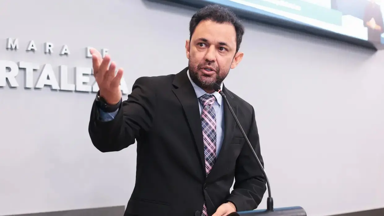 Homem de terno escuro e gravata fala ao microfone em um púlpito, gesticulando com a mão direita durante um discurso. Ao fundo, há uma parede clara com letras que indicam um ambiente institucional, possivelmente uma câmara legislativa.