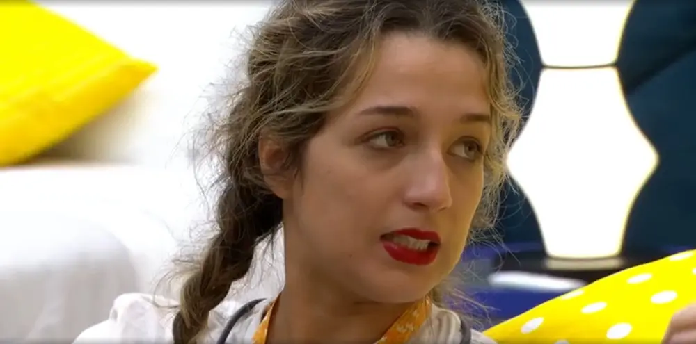 Na imagem, fotografia em close-focado de Samira, participante do BBB26, em um ambiente interno colorido. Ela tem pele clara, cabelos castanhos ondulados presos em uma trança lateral despojada e usa um batom vermelho vibrante. Sua expressão é séria e atenta, com o olhar direcionado para o lado. Ao fundo, veem-se elementos decorativos da casa, como almofadas amarelas e uma luminária branca com design geométrico.