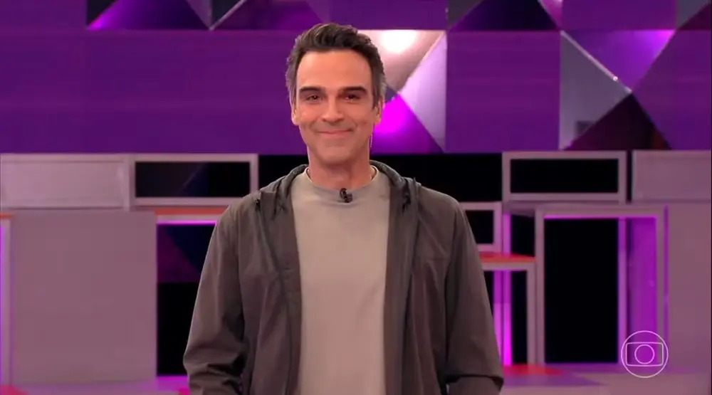 Na imagem, o apresentador Tadeu Schmidt aparece de frente, do peito para cima, sorrindo gentilmente para a câmera. Ele tem pele clara, cabelos escuros penteados para trás e veste uma camiseta cinza sob uma jaqueta aberta de tom cinza-escuro. O cenário ao fundo é o estúdio do BBB, composto por painéis geométricos em tons de roxo, lilás e preto, com iluminação moderna. No canto inferior direito, vê-se o logotipo da TV Globo.