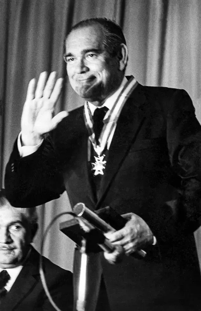 Na imagem, fotografia em preto e branco de Edson Queiroz, um homem de meia-idade, sorrindo e acenando com a mão direita levantada. Ele veste um terno escuro, camisa clara, gravata e carrega uma medalha de honra pendurada no pescoço por uma fita. Na mão esquerda, ele segura o que parece ser um estojo de premiação próximo a um microfone. Ao fundo, de forma parcial e desfocada, vê-se outro homem sentado e uma cortina de tecido pregueado.
