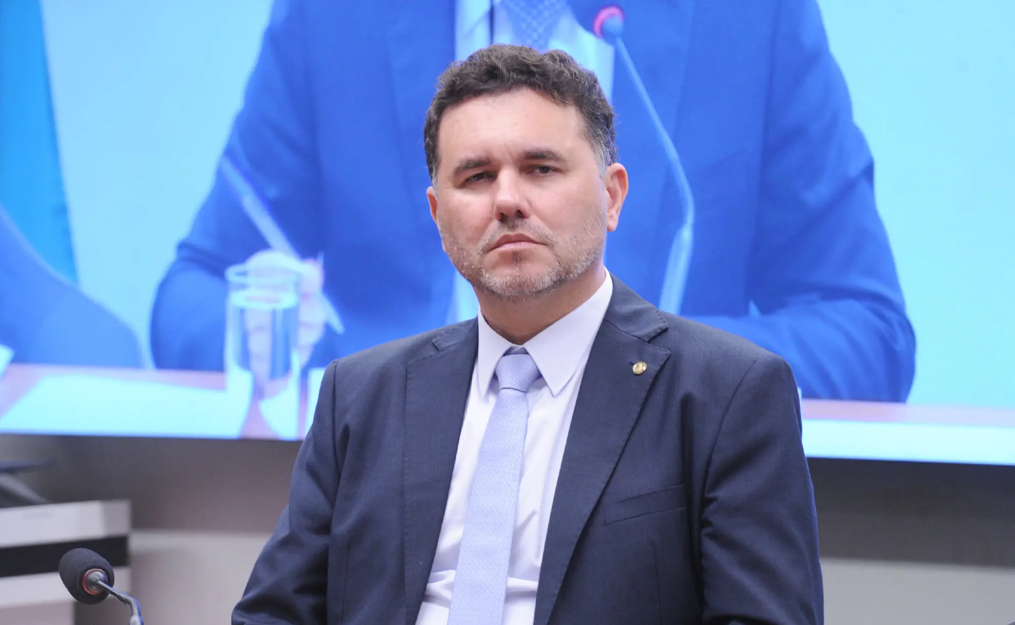 Homem de meia-idade, com expressão séria, sentado em um ambiente formal que parece ser uma audiência ou reunião oficial. Ele veste terno escuro, camisa branca e gravata clara, e possui barba curta e cabelo escuro levemente grisalho. À sua frente há um microfone sobre a mesa, e ao fundo aparece uma tela grande exibindo a imagem de outra pessoa falando ao microfone.