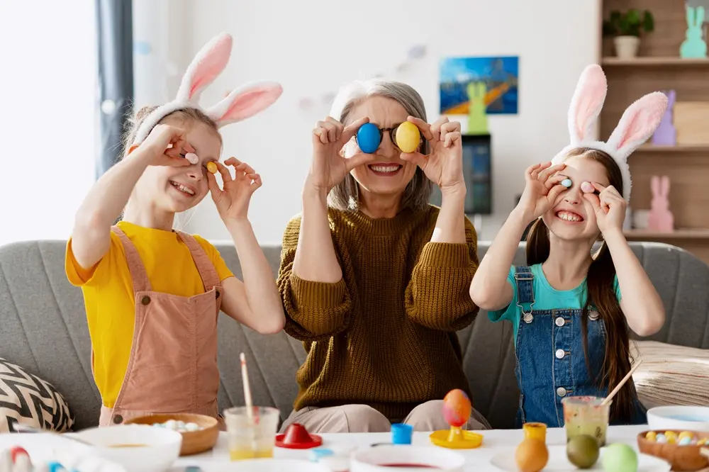Na imagem, uma mulher idosa e duas meninas sentadas à mesa em um momento descontraído de Páscoa. Elas usam orelhas de coelho e brincam de esconder os olhos com ovos coloridos. A menina à esquerda veste uma blusa amarela e jardineira bege; a mulher ao centro veste um suéter oliva; e a menina à direita veste uma blusa azul e jardineira jeans. À frente delas, sobre a mesa, há tintas, pincéis e ovos de Páscoa em diversas cores. O ambiente é iluminado e acolhedor.
