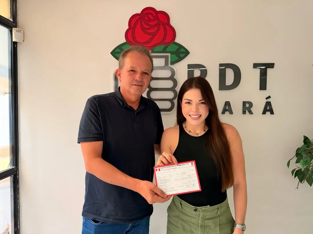 Homem e mulher posam lado a lado em ambiente interno, segurando um documento, em frente a um painel com o símbolo e a sigla de um partido político.