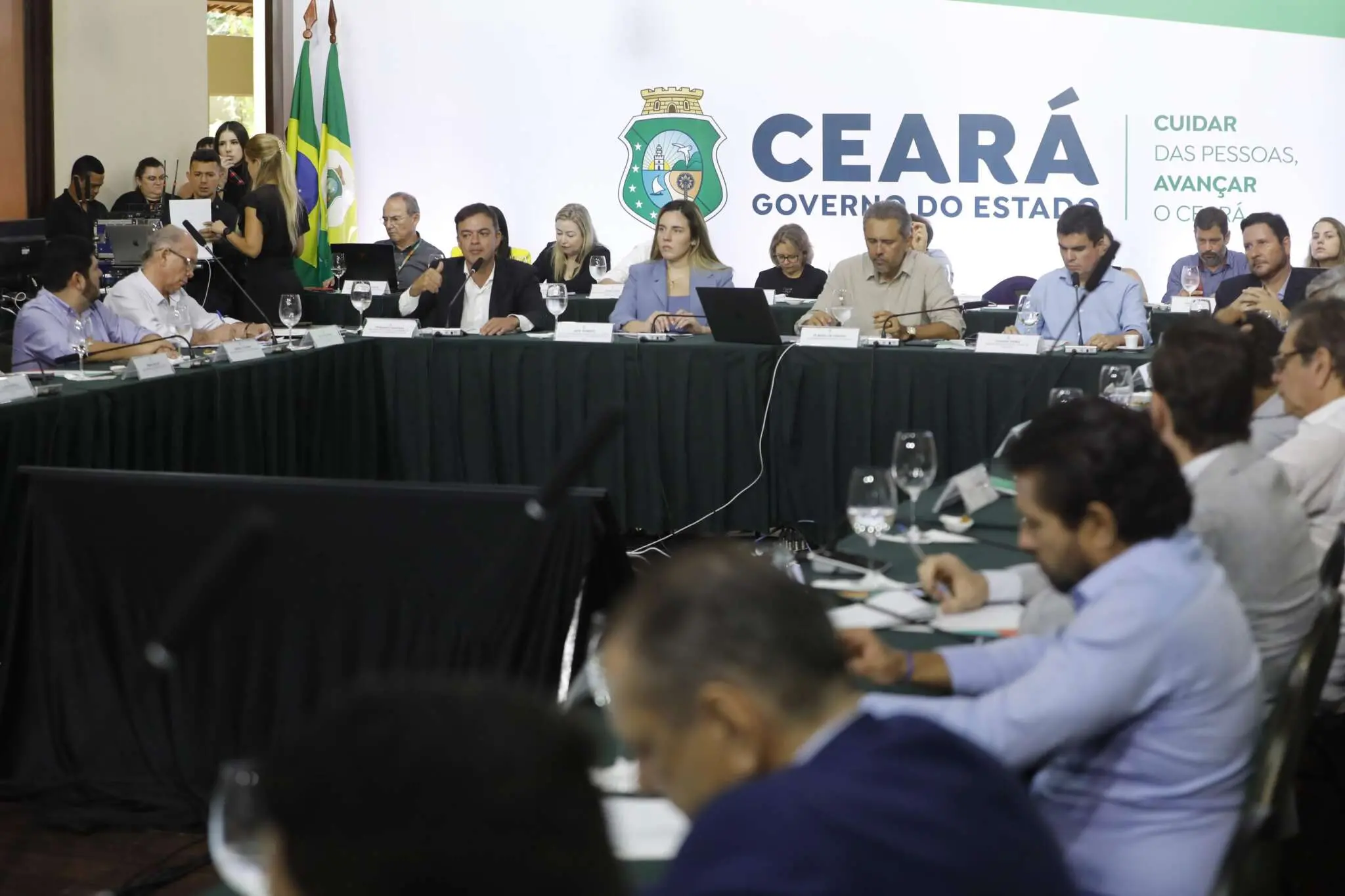 A imagem mostra uma reunião oficial do governo do estado do Ceará. Várias pessoas estão sentadas ao redor de uma mesa grande em formato de U, com laptops, microfones e copos de água à frente. No centro, um homem fala ao microfone enquanto os demais participantes escutam ou fazem anotações. Ao fundo, há uma parede com o logotipo e a inscrição “Ceará Governo do Estado”, além de bandeiras do Brasil e do estado. O ambiente é formal, com equipe de apoio e equipamentos técnicos visíveis ao lado.