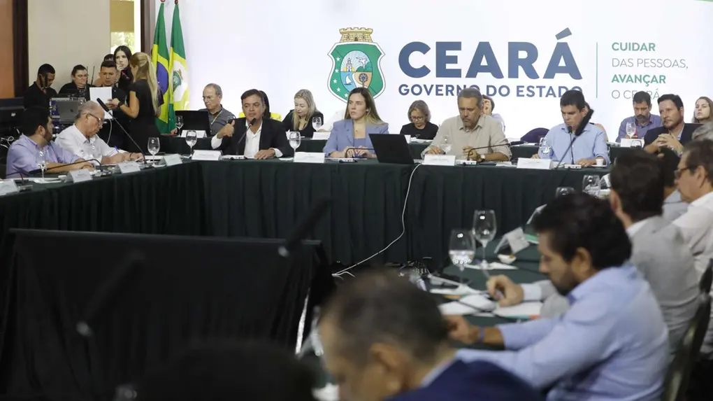 A imagem mostra uma reunião oficial do governo do estado do Ceará. Várias pessoas estão sentadas ao redor de uma mesa grande em formato de U, com laptops, microfones e copos de água à frente. No centro, um homem fala ao microfone enquanto os demais participantes escutam ou fazem anotações. Ao fundo, há uma parede com o logotipo e a inscrição “Ceará Governo do Estado”, além de bandeiras do Brasil e do estado. O ambiente é formal, com equipe de apoio e equipamentos técnicos visíveis ao lado.