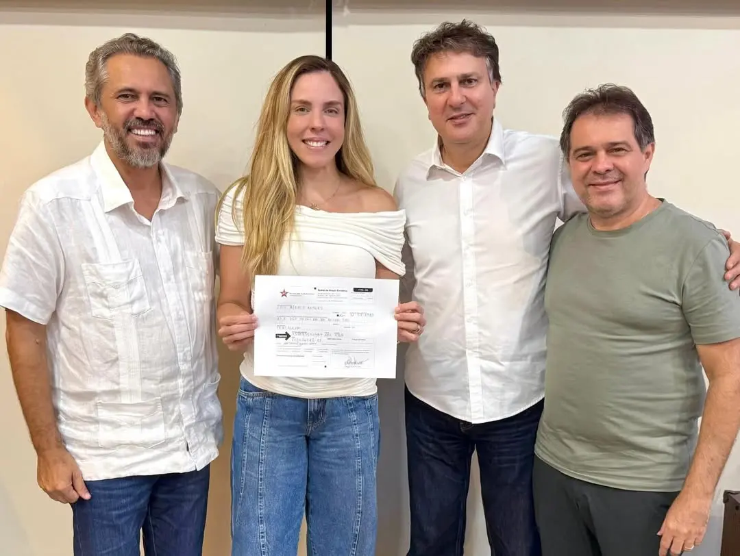 Quatro pessoas estão lado a lado, em um ambiente interno com parede clara ao fundo. No centro, uma mulher segura um documento com as duas mãos e sorri para a câmera. Ela veste uma blusa branca de ombro a ombro e calça jeans. Ao lado dela, há três homens também sorrindo: dois vestem camisas claras e um veste camiseta verde. Todos estão próximos, posando para a foto de forma informal.