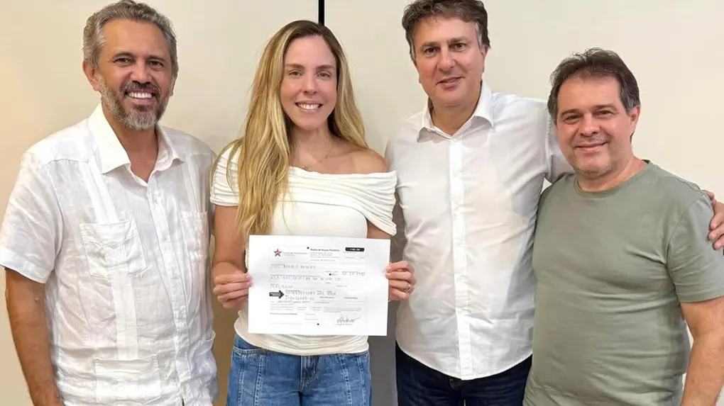 Quatro pessoas estão lado a lado, em um ambiente interno com parede clara ao fundo. No centro, uma mulher segura um documento com as duas mãos e sorri para a câmera. Ela veste uma blusa branca de ombro a ombro e calça jeans. Ao lado dela, há três homens também sorrindo: dois vestem camisas claras e um veste camiseta verde. Todos estão próximos, posando para a foto de forma informal.