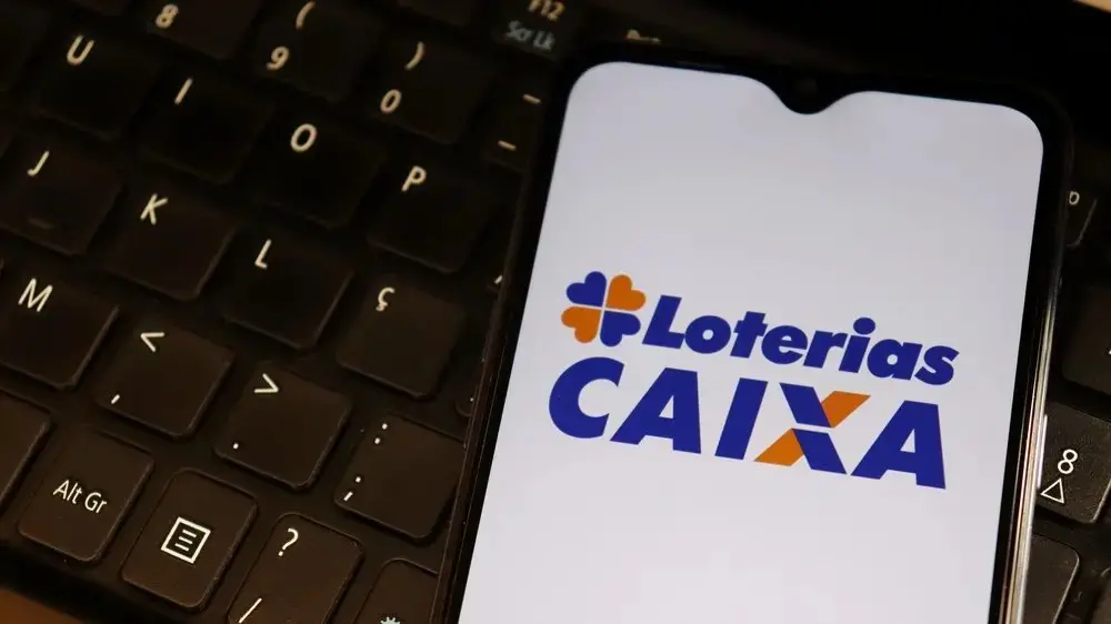 Imagem de um smartphone sobre o teclado de um computador exibindo na tela o logotipo “Loterias CAIXA” em fundo branco. O aparelho está inclinado e ocupa o centro da imagem, enquanto o teclado escuro aparece desfocado ao fundo.