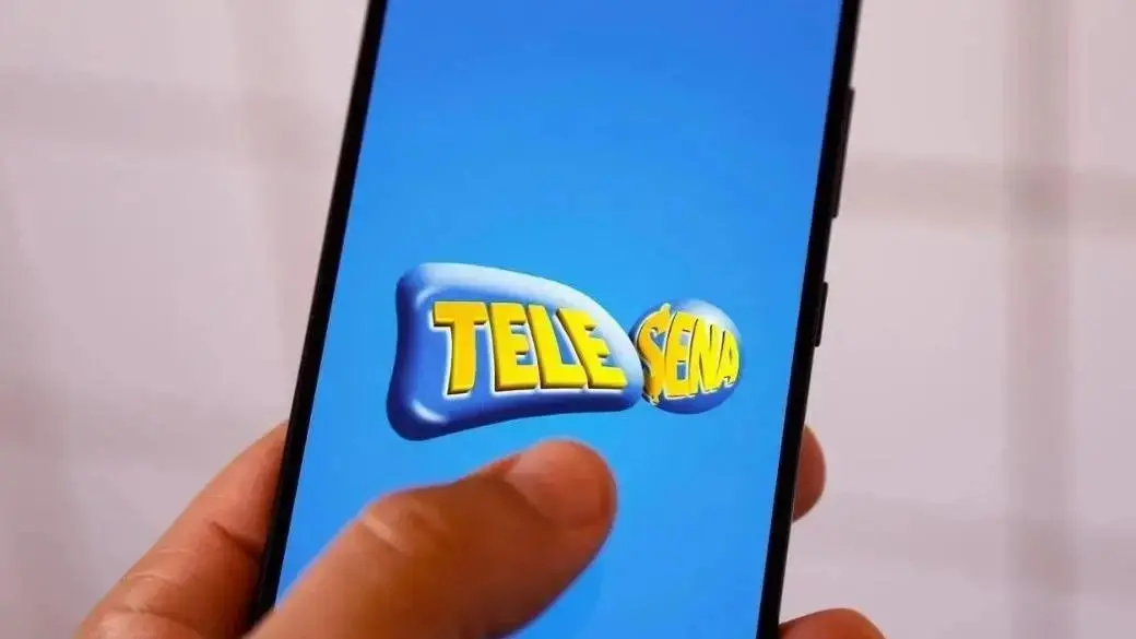 Pessoa segura um smartphone com a tela exibindo o logo azul e amarelo do Tele Sena, com o dedo prestes a tocar na tela.