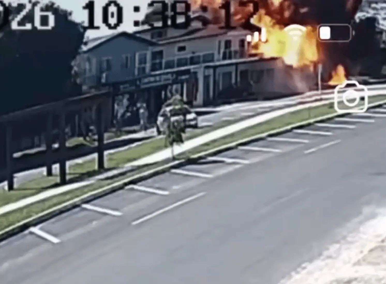 Imagem de câmera de segurança mostra o momento de uma explosão em um prédio comercial após a queda de uma aeronave. Uma grande bola de fogo aparece na parte superior direita do edifício, próximo ao telhado. Em frente ao local há uma rua com vagas de estacionamento vazias e uma calçada com árvores pequenas. A imagem tem data e hora no canto superior esquerdo e ícones de gravação no canto superior direito.