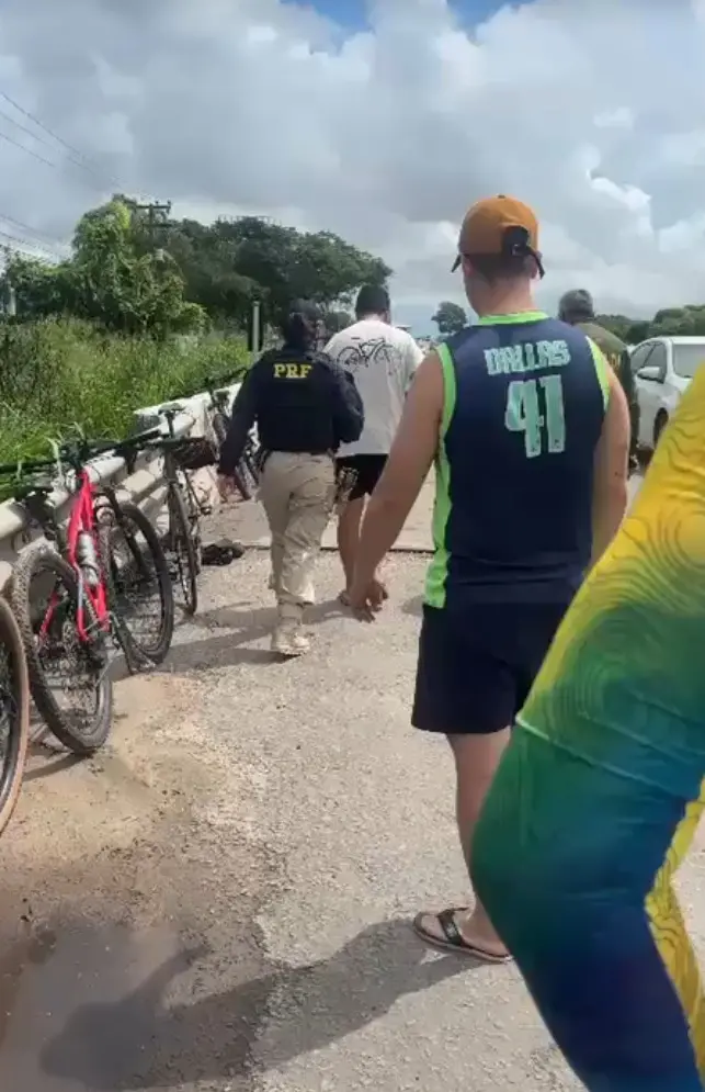Policial da PRF escolta pessoa na rodovia BR 116, em Itaitinga, no Ceará, enquanto bicicletas permanecem encostadas na estrada. O homem escoltado bateu em um grupo de ciclistas e foi preso por embriaguez.