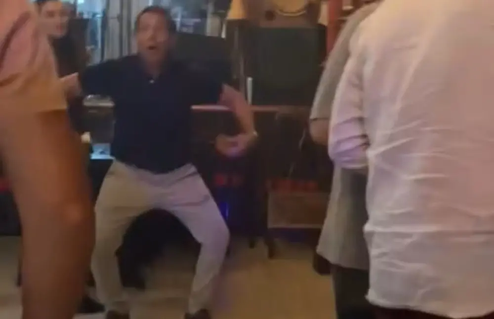 Na imagem, foto de baixa resolução capturada de um vídeo em um ambiente interno, possivelmente um restaurante ou bar. No centro, um homem de pele clara, vestindo uma camisa polo azul-marinho e calças claras, está em uma pose curvada com os joelhos dobrados e braços abertos para os lados. Ele tem uma expressão facial de surpresa ou exclamação, com a boca aberta. Outras pessoas aparecem parcialmente desfocadas em primeiro plano e ao fundo, sugerindo um momento de movimento ou interação social.