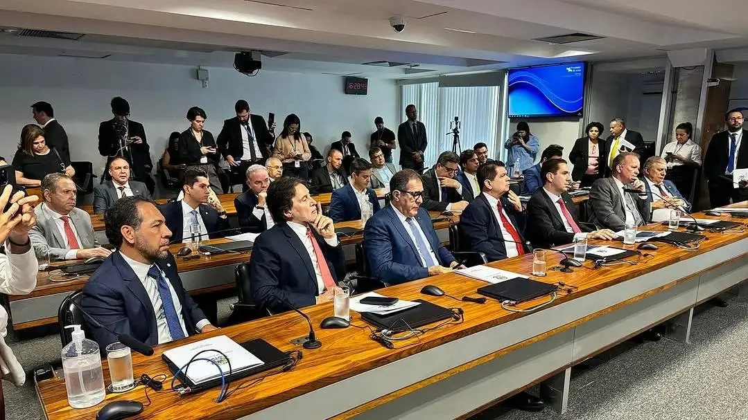 Sessão em sala de comissão com vários homens de terno sentados em longas mesas, usando microfones e laptops, enquanto outras pessoas acompanham ao fundo; ambiente formal de reunião política.