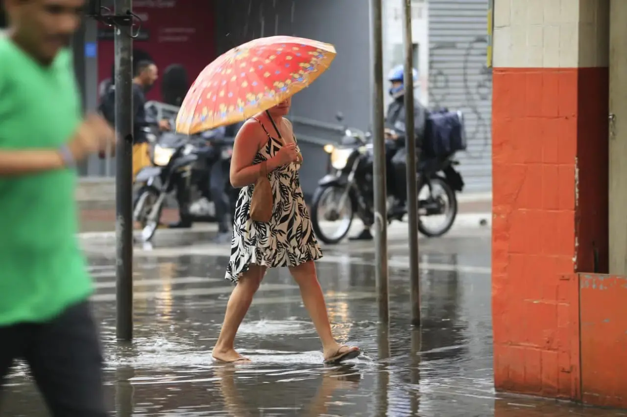 Uma mulher caminha sob chuva intensa em uma rua alagada, protegendo-se com um guarda-chuva estampado em tons de vermelho e laranja. Ela veste um vestido curto de folhagens em preto e branco e chinelos, enquanto ao fundo, motociclistas aguardam no asfalto molhado que reflete a luz da cidade.