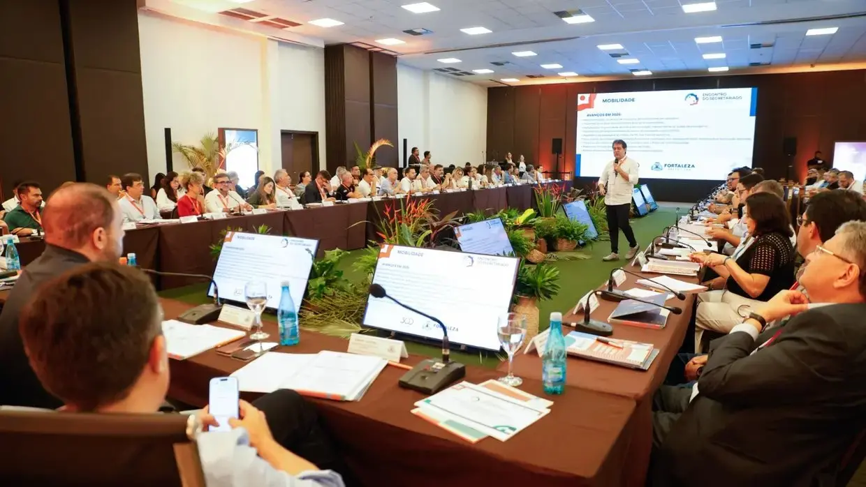 Reunião de secretariado em sala ampla, com participantes sentados em mesas em formato de U, enquanto um homem apresenta tema de mobilidade em frente a um telão; notebooks, microfones e materiais de trabalho sobre as mesas.