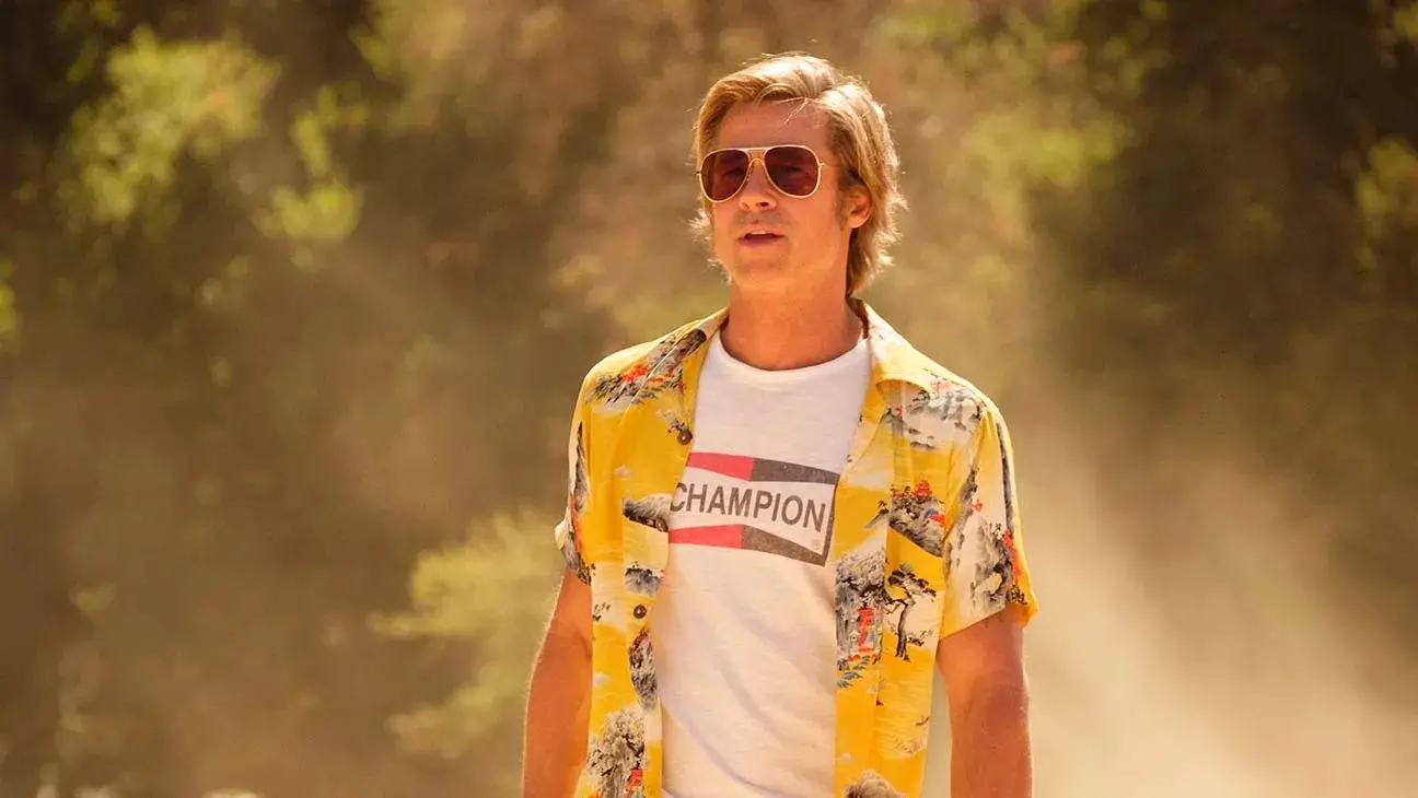 Brad Pitt aparece como o personagem Cliff Booth, vestindo uma camisa amarela estampada sobre uma camiseta branca com a logomarca 'Champion' e óculos de sol clássicos. Ele caminha em um ambiente ensolarado e levemente empoeirado, com uma vegetação desfocada ao fundo que reforça a estética vintage e cinematográfica da cena.