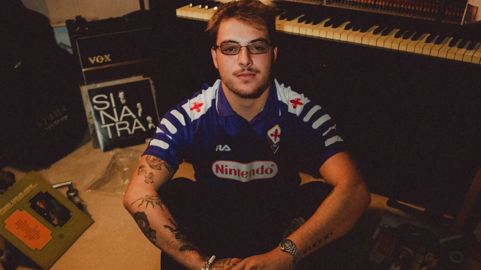 Homem usando camisa da Fiorentina com patrocínio da Nintendo aparece sentado em estúdio de música, cercado por teclado, amplificador Vox e discos de vinil, em foto de ambiente artístico. O homem na foto é o filho de Patrícia Poeta, Felipe Poeta, que celebrou melhora após 19 cirurgias.