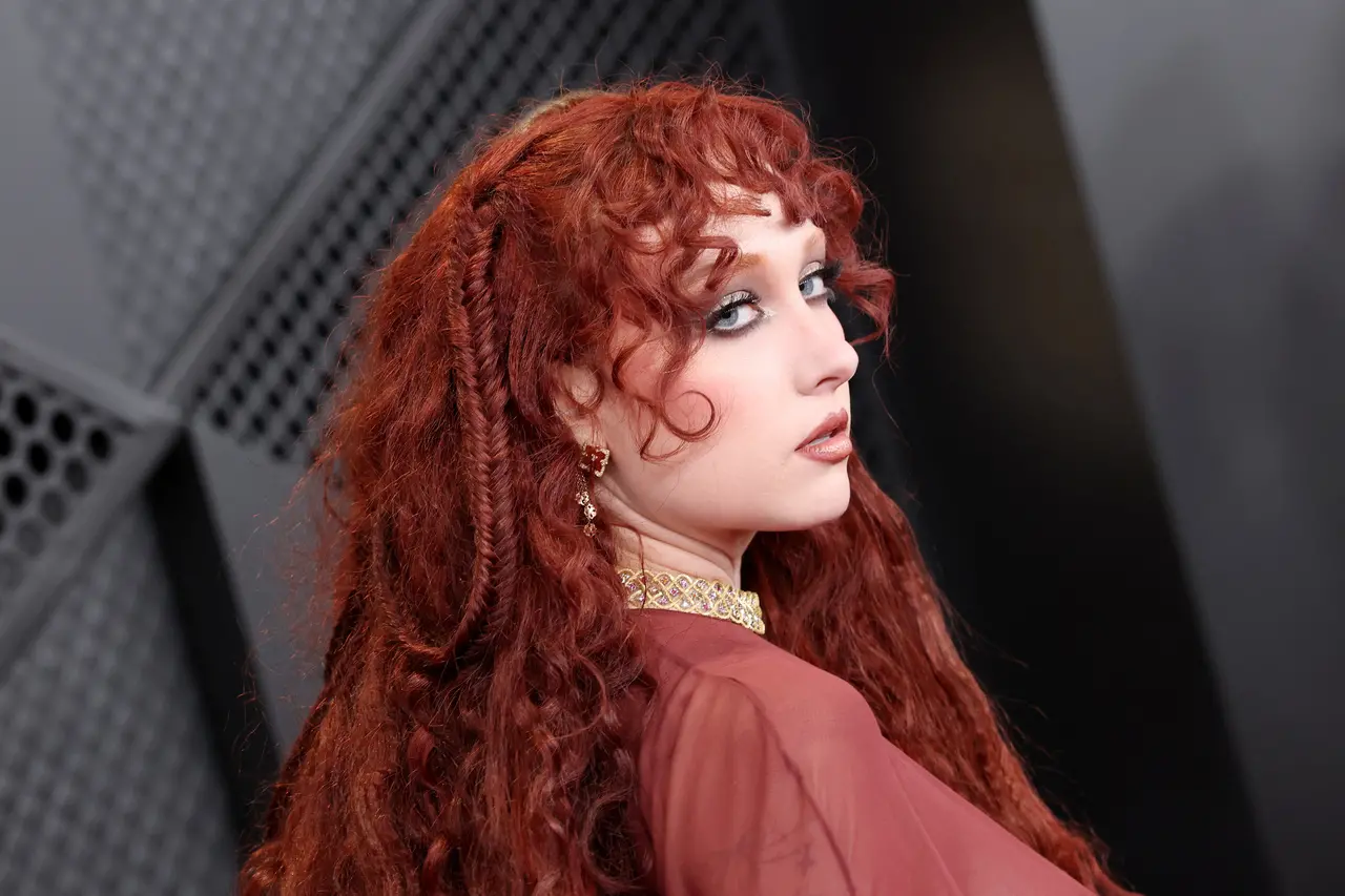 A cantora Chappell Roan posa de perfil exibindo seus longos cabelos ruivos e ondulados, que incluem pequenas tranças e uma franja encaracolada. Ela usa uma maquiagem marcante com tons prateados nos olhos, joias douradas e um traje em tom bordô que harmoniza com a paleta de cores vibrante da imagem.