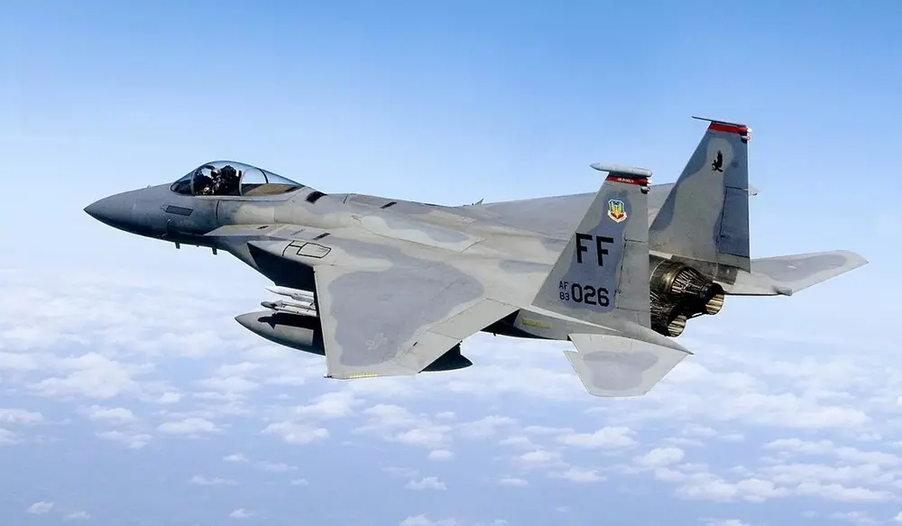 Na imagem, vista aérea de um caça F-15 Eagle em pleno voo sobre uma camada de nuvens brancas sob um céu azul claro. A aeronave militar possui pintura de camuflagem em tons de cinza e exibe a inscrição 