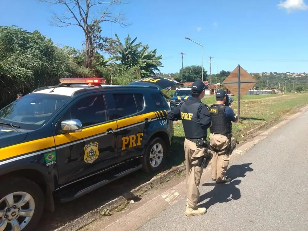 Na imagem, fotografia de uma operação da Polícia Rodoviária Federal (PRF) em uma estrada sob luz solar intensa. Em primeiro plano, à esquerda, está estacionada uma viatura ostensiva da PRF, um SUV azul-marinho com faixas amarelas e o giroflex ligado. À direita da viatura, dois policiais fardados com calças cáqui e coletes pretos com a inscrição 