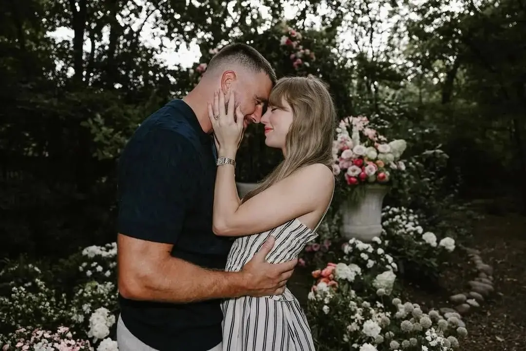 Casal abraçado em um jardim decorado com flores. O homem segura a mulher pela cintura enquanto ela toca o rosto dele, e os dois encostam a testa um no outro, sorrindo. Ao fundo, há arranjos florais em tons de branco, rosa e vermelho, além de árvores e folhagens, criando um ambiente romântico ao ar livre.
