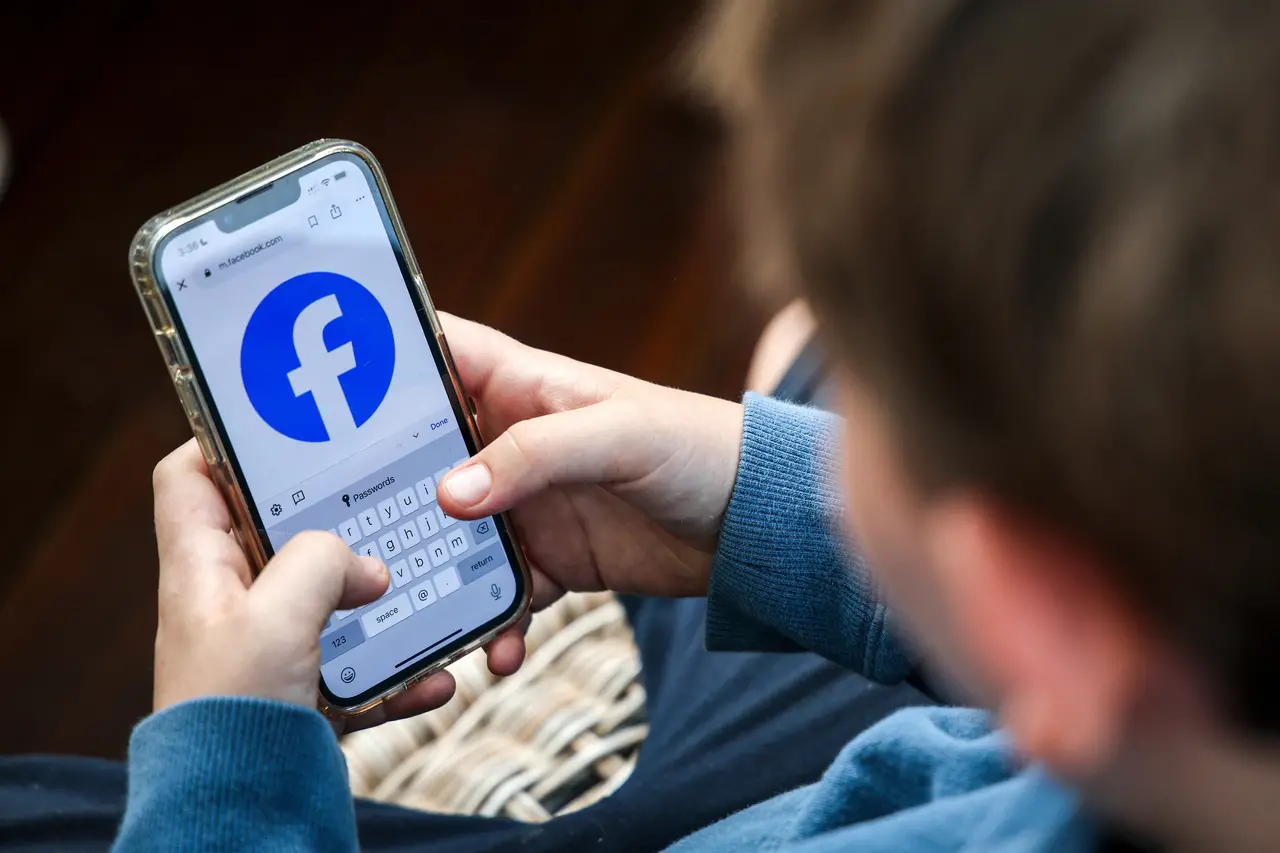 Uma criança segura um smartphone com as duas mãos, exibindo a página de login do Facebook com o logotipo azul em destaque na tela. O plano fechado foca nos dedos da criança sobre o teclado virtual do aparelho, enquanto o fundo permanece levemente desfocado.