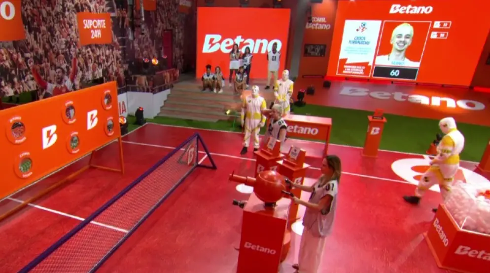 Quadra cenográfica de prova com piso vermelho e elementos nas cores laranja e branco, com a marca “Betano” em destaque nas paredes e telões. Em primeiro plano, uma participante veste uniforme branco e segura uma alavanca ligada a um equipamento esférico vermelho. À esquerda, há uma pequena rede semelhante à de tênis e um painel com alvos circulares. Ao fundo, outras pessoas com uniformes brancos e amarelos aguardam, enquanto um telão exibe a foto de um participante e o número 60. O cenário lembra uma competição de programa de televisão.