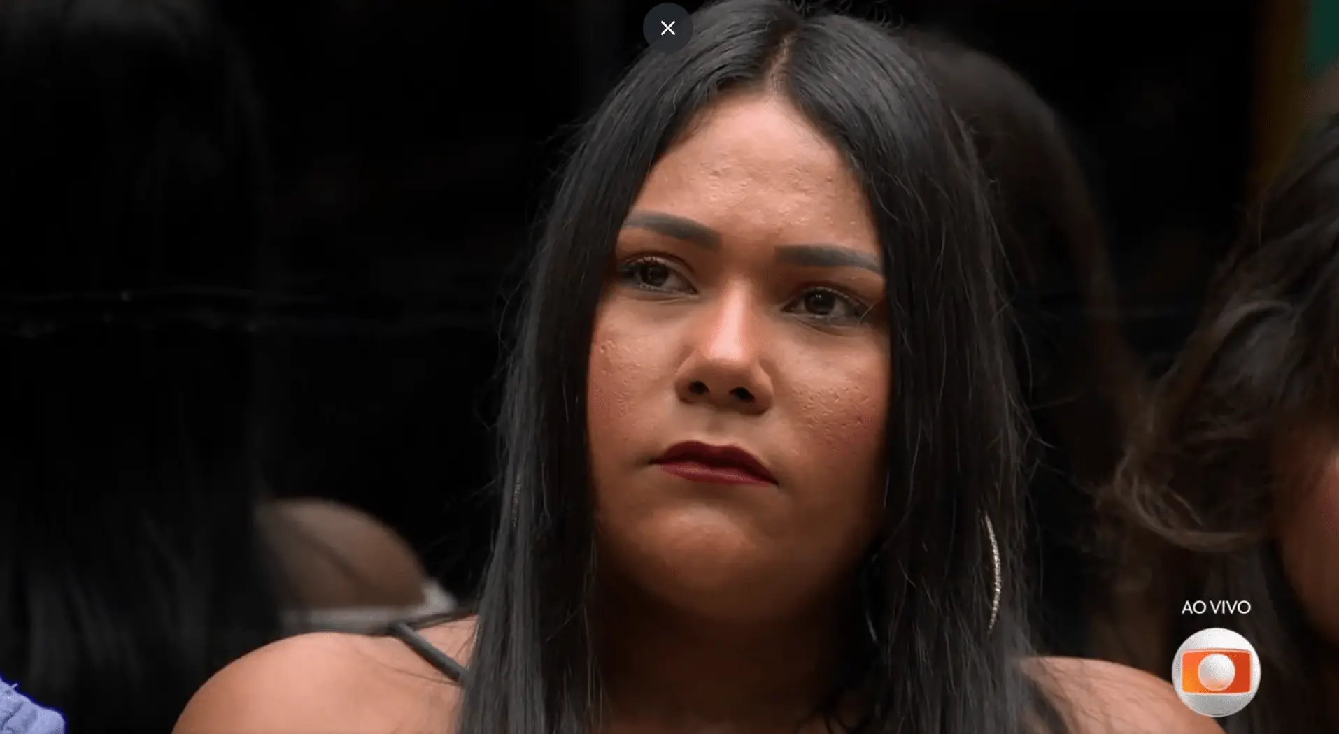Mulher de cabelos longos e pretos, usando brincos de argola e blusa de alça, aparece em close com expressão séria e olhar fixo à frente. A imagem parece ser de um programa de televisão ao vivo, com o logotipo da TV Globo e a indicação “AO VIVO” no canto inferior direito. O fundo está escuro e desfocado, com outras pessoas parcialmente visíveis ao redor.
