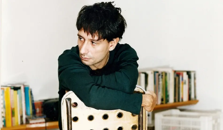 O músico Alvin L aparece sentado de costas em uma cadeira branca com furos, apoiando os braços no encosto enquanto olha pensativo para o lado. Ao fundo, uma estante repleta de livros coloridos e uma parede branca completam o cenário íntimo e casual.