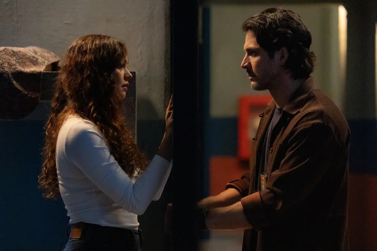 Duas pessoas se encaram em um ambiente interno pouco iluminado, separadas por uma parede; uma apoia a mão na superfície enquanto a outra mantém os braços cruzados, sugerindo uma conversa tensa ou íntima.