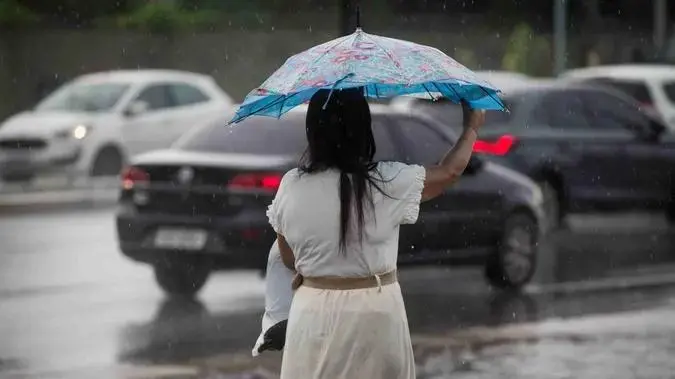 Inmet recomenda cuidados para evitar acidentes. Mulher se protege de chuva usando guarda-chuva. Ao fundo, é possível ver carros trafegando em uma via.