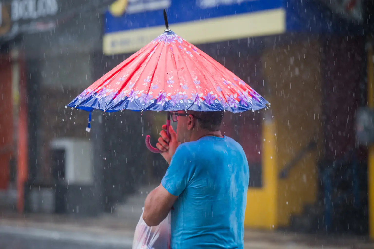Homem de costas sob chuva moderada, vestindo camiseta azul e segurando uma sacola plástica branca. Ele se protege com um guarda-chuva colorido de estilo oriental, com tons de vermelho, azul e estampas florais. O fundo urbano está desfocado, destacando as gotas de chuva caindo.
