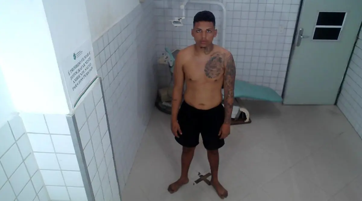 Homem jovem, de pé, em uma sala com paredes e piso revestidos de azulejos claros, aparentando ambiente institucional. Ele está sem camisa, vestindo bermuda preta e descalço. O homem tem cabelo curto e tatuagens no braço esquerdo e no lado esquerdo do peito. No fundo da sala, há uma maca encostada na parede e uma porta à direita.