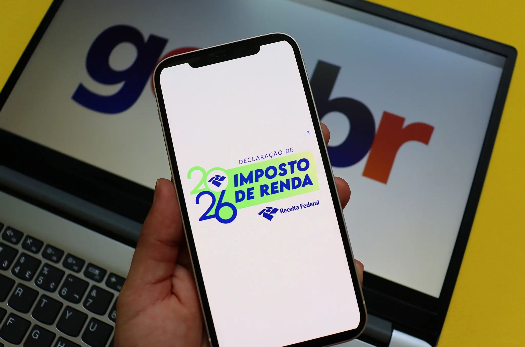 Logo da Declaração do Imposto de Renda 2026 exibida em um celular. Uma tela de computador, ao fundo, mostra o logotipo Gov.br.