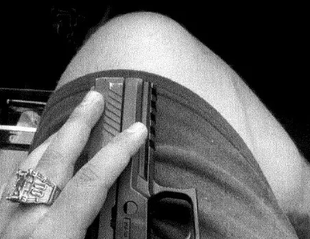 Foto em preto e branco de um dos suspeitos ostentando arma da Polícia Militar. Não aparece o rosto dele, só a mão e uma parte da perna, além da arma.