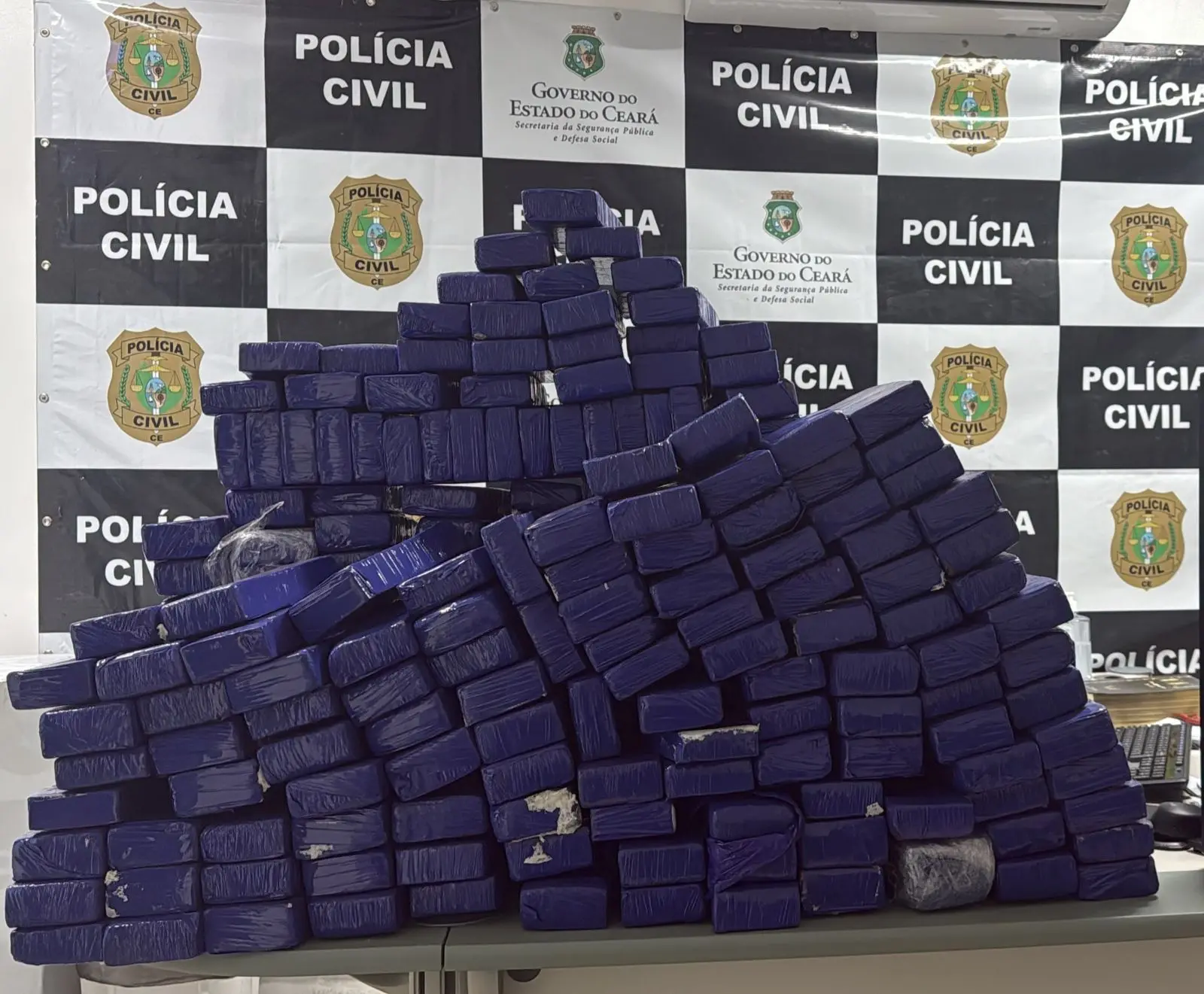 Grande quantidade de drogas apreendidas pela Polícia Civil do Ceará, exibindo blocos empilhados em uma mesa com fundo de painel de polícia.