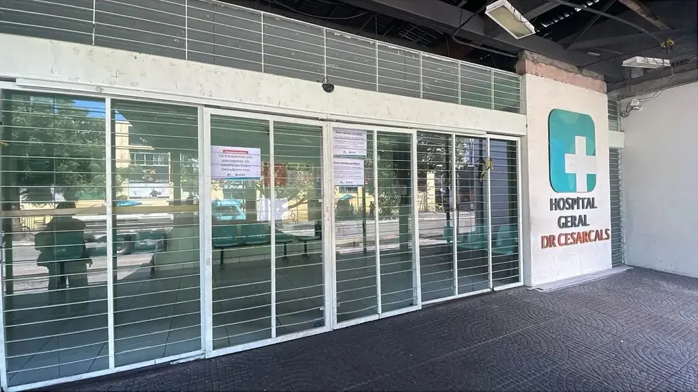 Fachada do Hospital Geral Dr. César Cals, em Fortaleza, com grades metálicas fechando a entrada principal. Há avisos impressos afixados nos vidros, e o interior aparenta estar vazio, com cadeiras alinhadas. O letreiro com o nome do hospital aparece à direita, ao lado do símbolo de cruz em verde.
