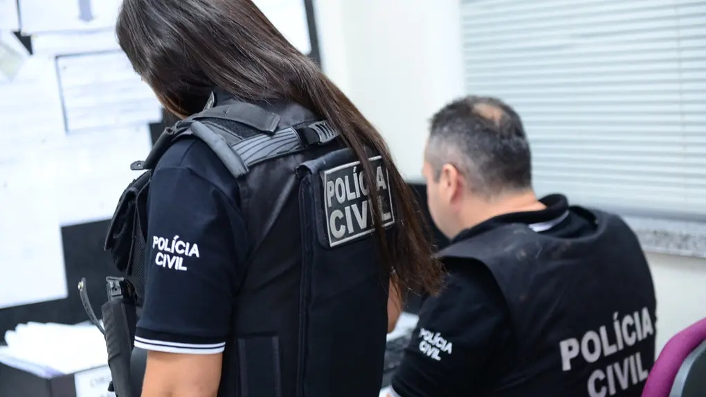 Policiais civis em ambiente de escritório. Em primeiro plano, uma agente de costas usa colete tático com a inscrição “Polícia Civil” e observa uma tela. Ao lado, um agente sentado também com uniforme da Polícia Civil trabalha em frente a um computador. O ambiente é claro, com mesas, papéis e equipamentos de informática visíveis.