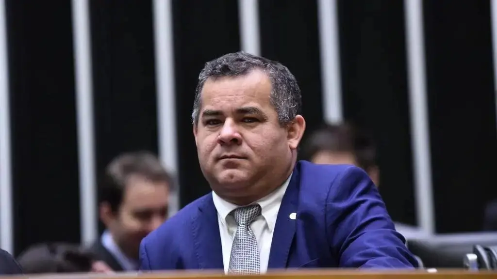 Homem de terno azul e gravata clara aparece sentado em plenário legislativo, com expressão séria, enquanto outras pessoas desfocadas estão ao fundo.