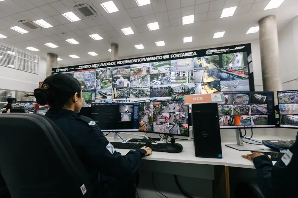 Na imagem, uma mulher de farda azul-marinho, vista de costas e de perfil, está sentada em uma estação de trabalho operando um centro de monitoramento. Ela observa diversos monitores à sua frente; em sua mesa, há dois monitores menores exibindo várias divisões de câmeras de segurança, um teclado e um mouse. Ao fundo, domina a cena um grande painel de vídeo (videowall) que ocupa quase toda a largura da parede. O painel exibe dezenas de transmissões ao vivo de câmeras de rua, mostrando avenidas, cruzamentos e espaços públicos. No topo do painel, lê-se o letreiro: 