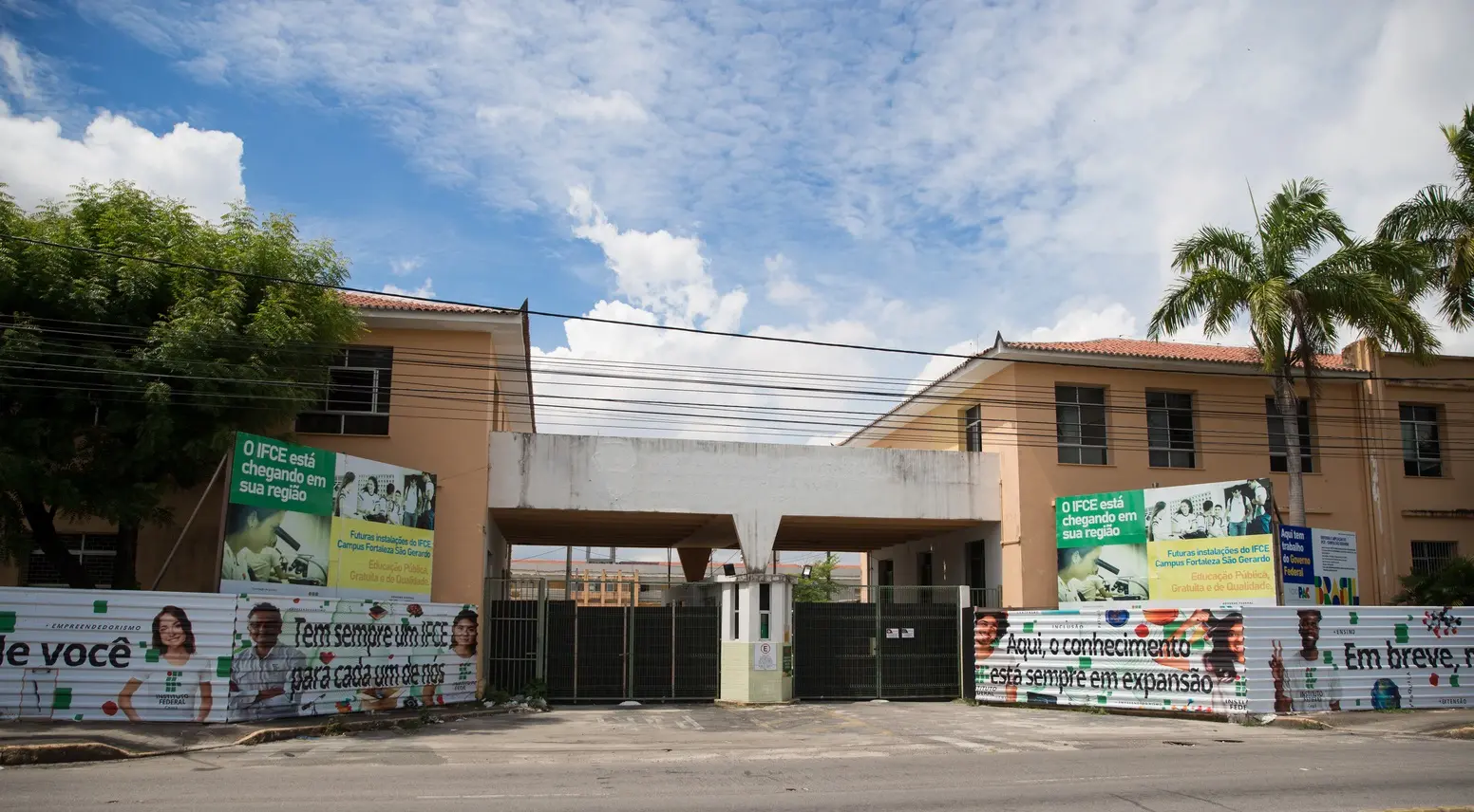 Vista frontal ampla da entrada principal do campus, composta por dois blocos simétricos unidos por um pórtico central de concreto, protegida por portões pretos e grandes painéis informativos do IFCE.