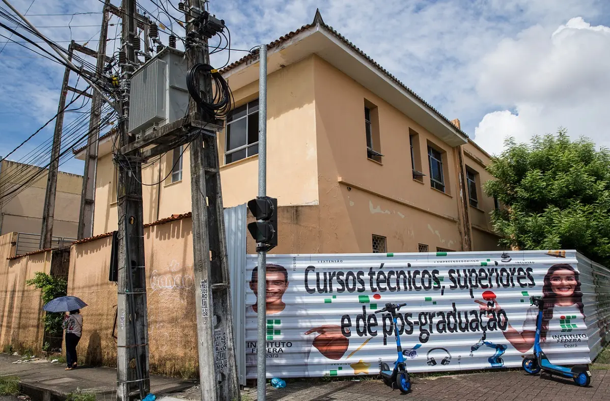 Detalhe da lateral do edifício focado nas esquadrias das janelas superiores, com um poste de transformação de energia à esquerda e patinetes elétricos azuis estacionados junto ao tapume.