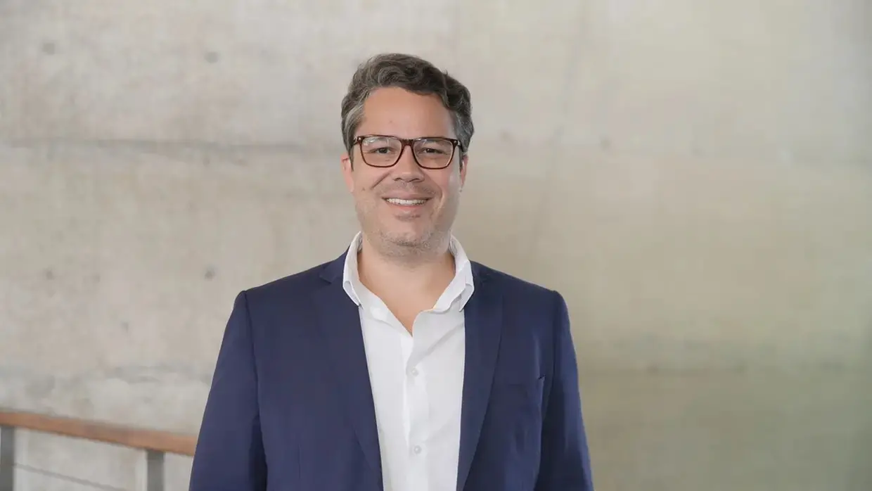 Homem de terno azul marinho e camisa branca em frente a fundo neutro.