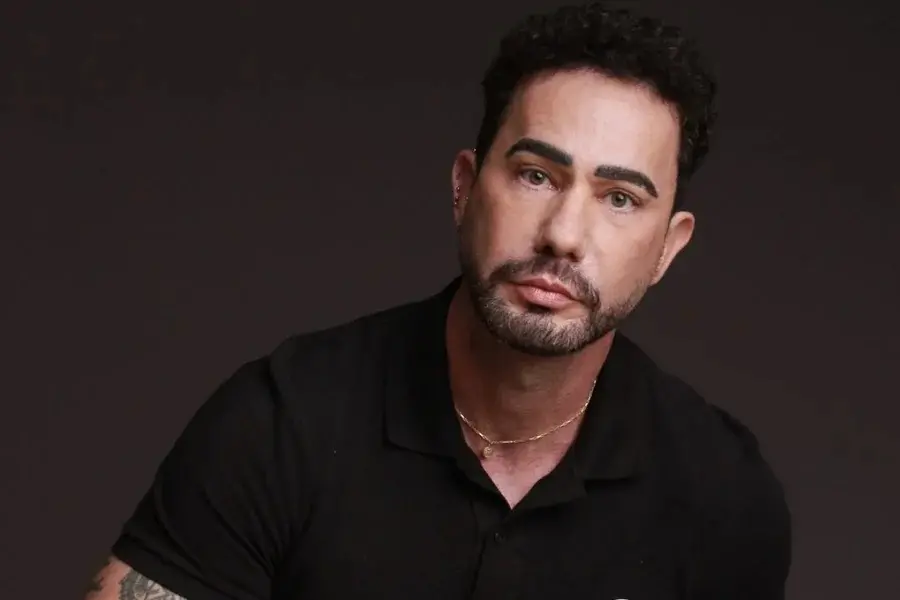 Homem de pele clara e barba aparada posa em um fundo escuro, vestindo uma camisa polo preta e uma corrente dourada fina. Ele encara a câmera com uma expressão séria, destacando o contraste entre a iluminação suave e as sombras que definem seus traços.