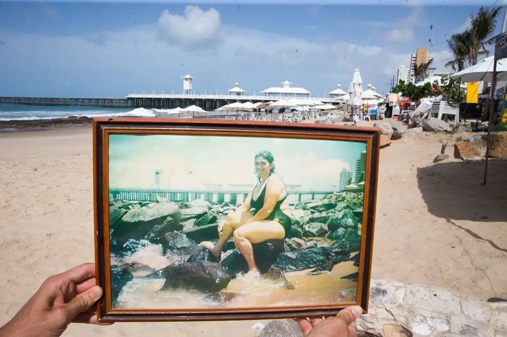Na imagem, uma fotografia em perspectiva que sobrepõe o passado e o presente na Ponte dos Ingleses, em Fortaleza. No primeiro plano, duas mãos seguram um quadro emoldurado contendo uma foto antiga, analógica e levemente desbotada. Na foto antiga (dentro do quadro): Uma mulher sorridente, vestindo um maiô preto, está sentada sobre rochas à beira-mar. Ao fundo, vê-se a estrutura original da ponte sob um céu claro. No plano de fundo (cenário real): A mesma praia e a estrutura atual da Ponte dos Ingleses (também conhecida como Ponte Metálica) se estendem pelo horizonte. A areia está ocupada por guarda-sóis brancos, e o céu apresenta nuvens esparsas.

A composição cria um contraste visual entre a memória afetiva preservada no quadro e a paisagem urbana contemporânea ao fundo.