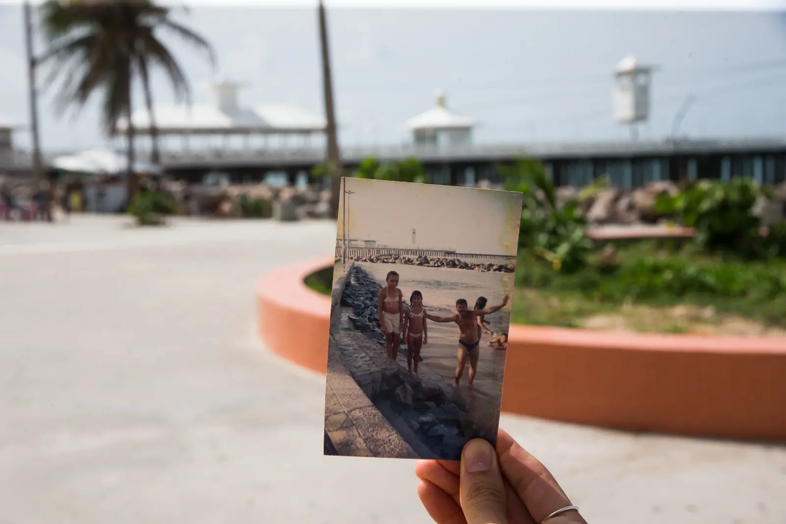 Na imagem, uma fotografia que utiliza a técnica de sobreposição para contrastar o passado e o presente na Praia de Iracema, em Fortaleza. Uma mão segura uma foto analógica vertical e desgastada pelo tempo, alinhando-a com a paisagem atual ao fundo. Na foto antiga (em destaque): Um homem e duas crianças em trajes de banho posam felizes em uma faixa de areia estreita, junto a uma mureta de pedras. Ao fundo da foto, vê-se a silhueta da Ponte dos Ingleses e o mar. No plano de fundo (cenário real): A área aparece revitalizada, com um amplo calçadão de concreto claro, um banco curvo pintado de terracota e vegetação rasteira. A estrutura da Ponte dos Ingleses, com seus quiosques brancos característicos, aparece ao fundo, levemente fora de foco. A imagem destaca as transformações urbanas da orla, mantendo viva a memória afetiva de quem frequenta o local há décadas.
