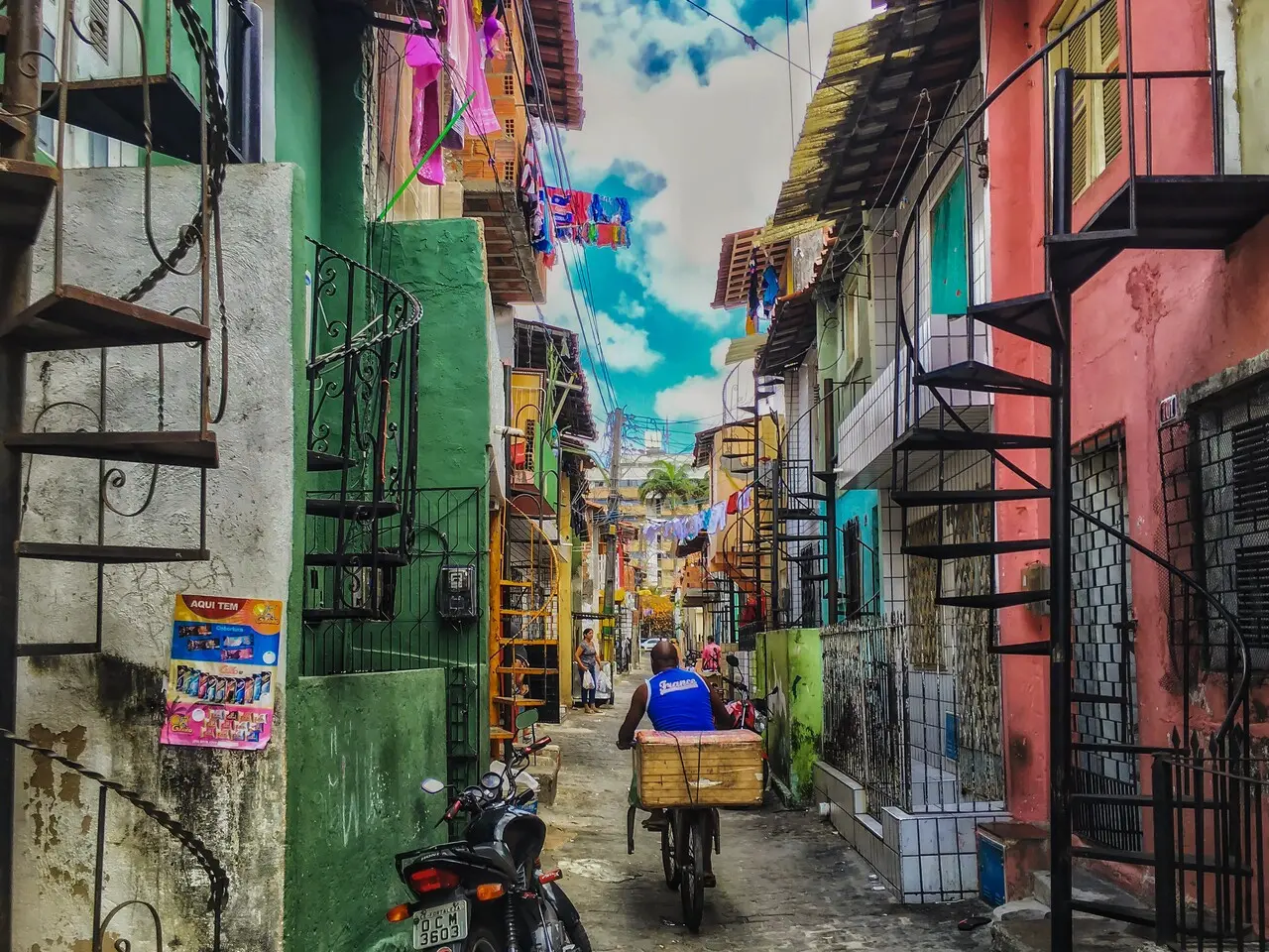 Um entregador em uma bicicleta de carga percorre um beco estreito cercado por casas coloridas e escadas caracol metálicas que sobem pelas fachadas. Roupas estendidas em varais e o céu azul com nuvens ao fundo compõem uma cena urbana vibrante e cotidiana.