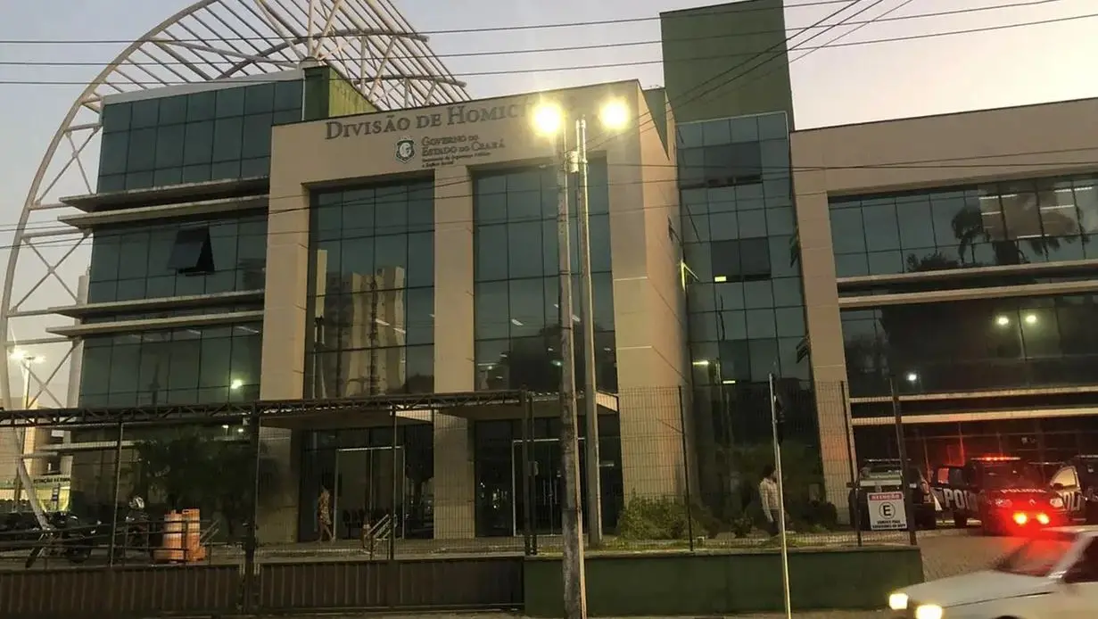 Fachada do prédio da Divisão de Homicídios da Polícia Civil do Ceará, com estrutura moderna de vidro, letreiro institucional na parte superior e viaturas estacionadas em frente ao edifício ao entardecer.